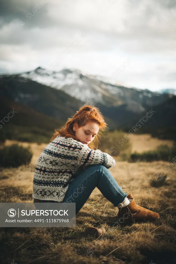 Side view of pensive charming woman with red hair in warm sweater sitting with closed eyes on lawn in mountain in gray day;Red haired woman thoughtfully sitting on meadow in mountains