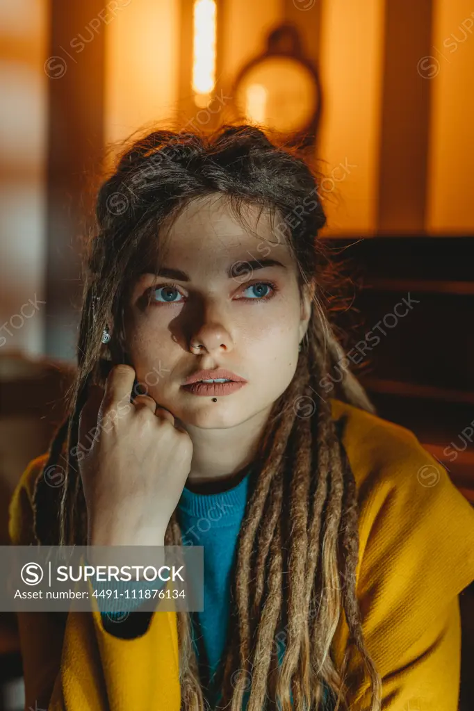 Stylish millennial female with dreadlocks wearing yellow coat over blue sweater looking away thoughtfully while sitting in room with piano in background;Pensive hipster woman looking away