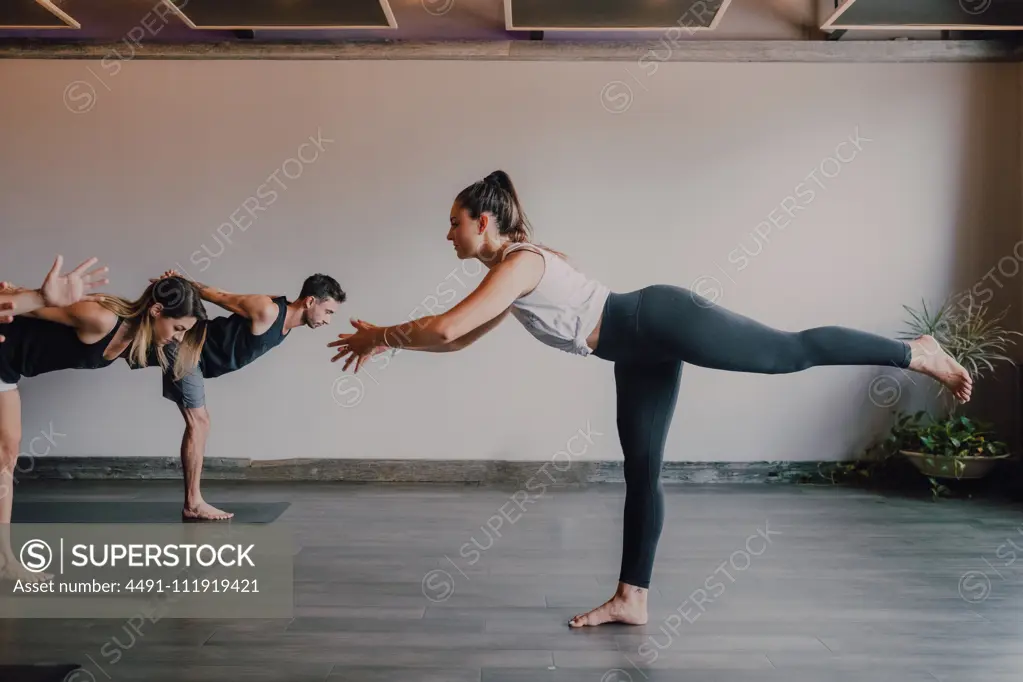 Slim barefoot female instructor in sportswear standing in warrior pose three and controlling group of sporty people concentrating and doing same exercise standing on sports mats in modern workout room