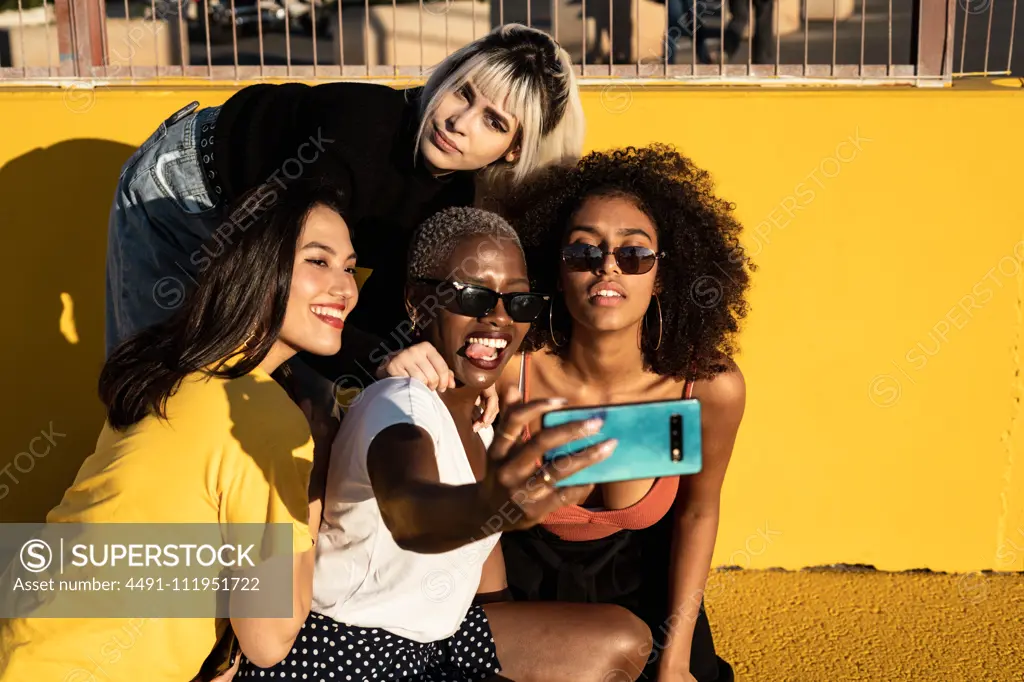 Cheerful young diverse female friends taking selfie on smartphone in street
