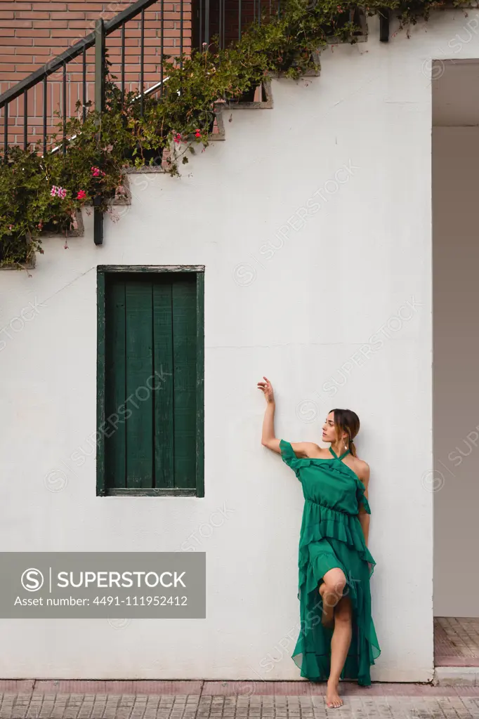 Full body barefoot female in green dress leaning on white wall of house with staircase and potted flowers on town street