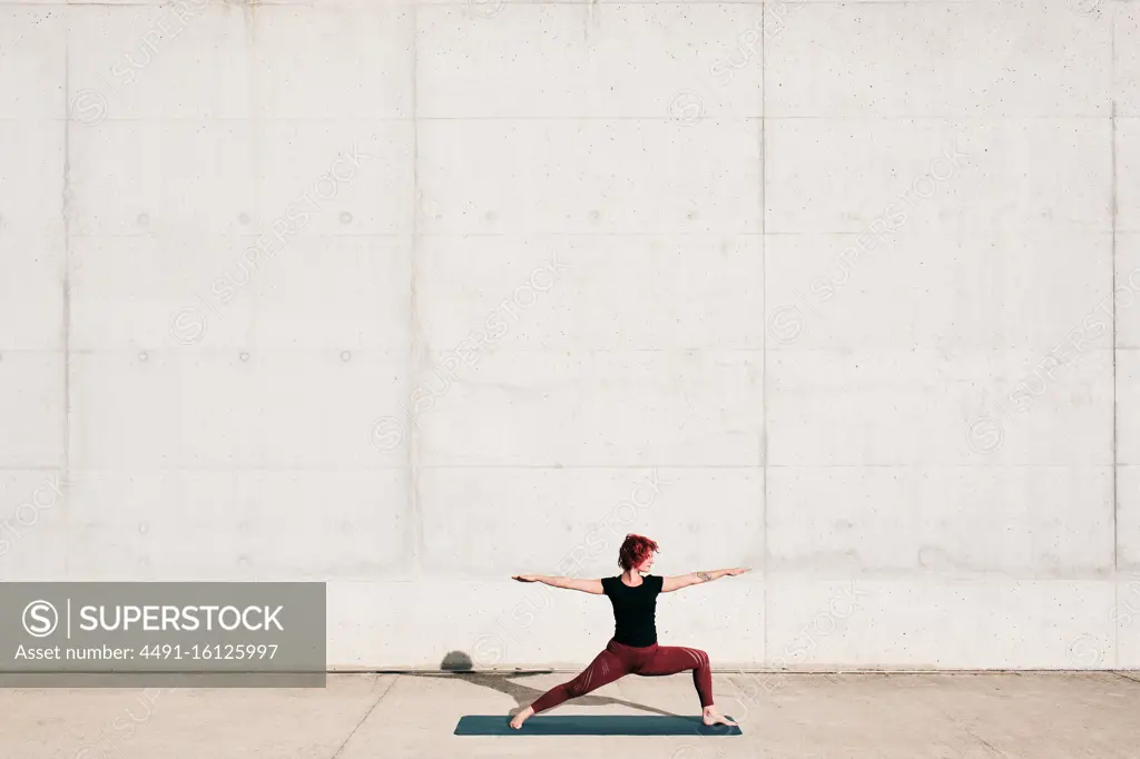 Side view of trendy fit female athlete in sportswear doing warrior two yoga pose on sports mat while training alone on street against concrete wall in sunny day