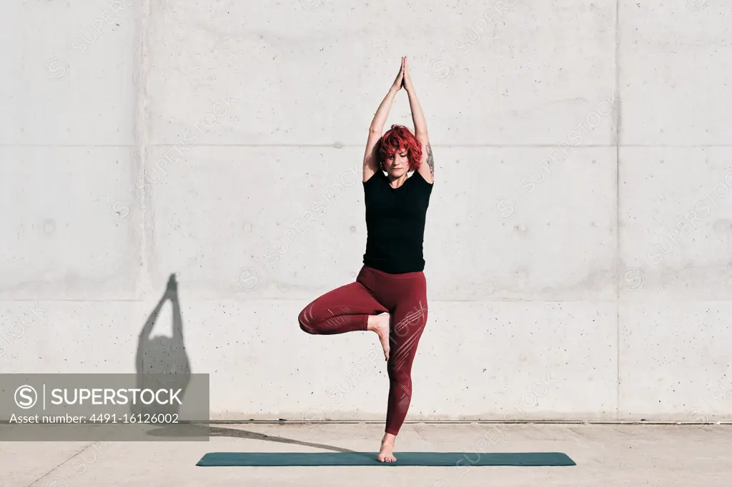 Confident barefooted female athlete in sportswear meditating while standing in vrikshasana position with arms raised and namaste on sports mat training alone on street against concrete wall in sunlight