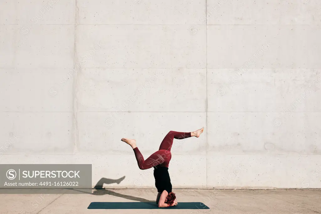 Side view of unrecognizable barefooted female athlete in activewear standing upside down in sirsasana position with legs raised in split on sports mat training alone on street against concrete wall in daytime