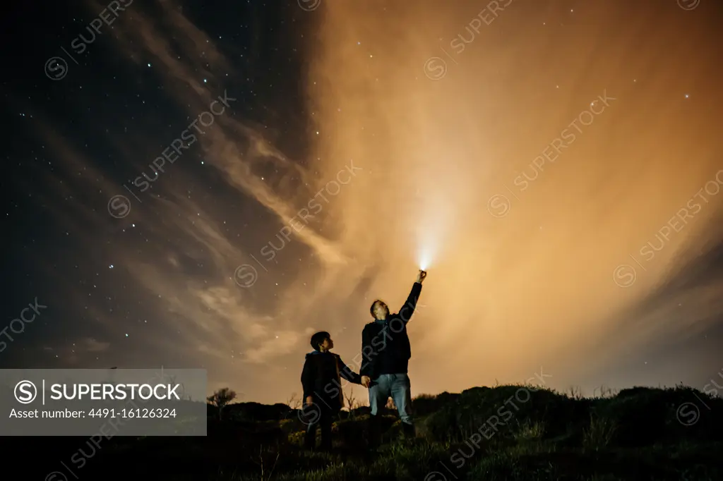 Carefree dad and son holding hands and observing starry sky while looking up and lighting with torch at night