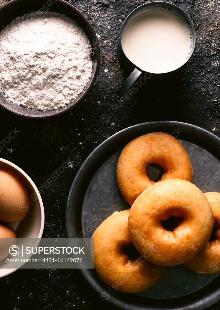 Top view of fresh doughnuts placed on rough table near various pastry ingredients and utensils in kitchen