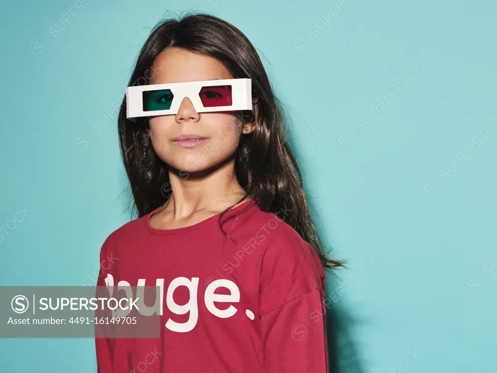 Happy preteen girl in white plastic stereoscopic glasses with red and blue lens smiling at camera while watching 3d graphic film with pleasure against turquoise background in modern studio