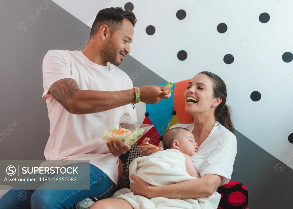 Mother with baby talking on mobile phone while sitting on armchair with casual father eating salad