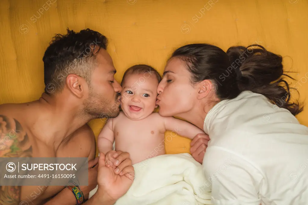 Top view of young happy parents kissing in cheeks little satisfied baby while lying on bed