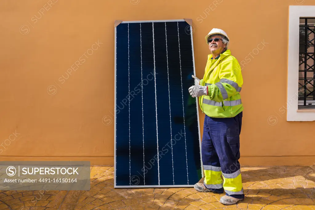 Side view of mature experienced male technician in uniform and helmet standing with solar panel near yellow building while working on installation of renewable energy system