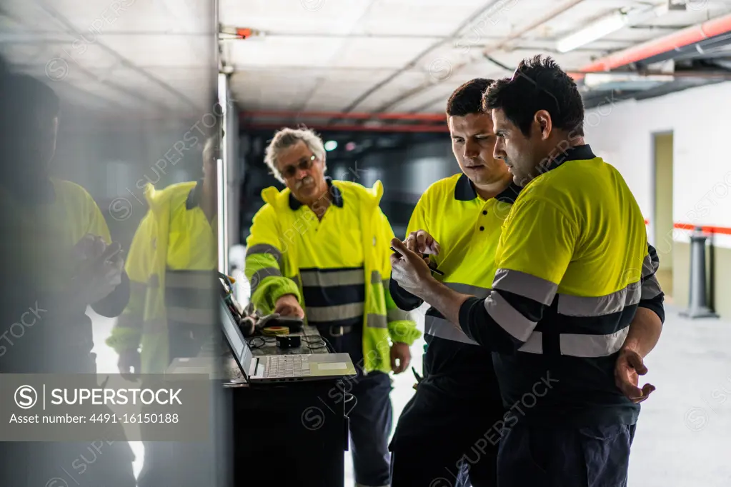 Group of skilled male engineers in uniform using gadgets while examining electrical equipment in modern building