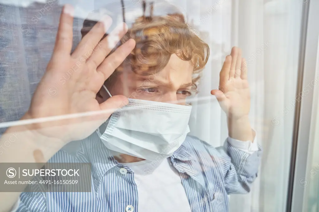 Boy in medical mask keeping hand on glass and looking through window while staying home during quarantine