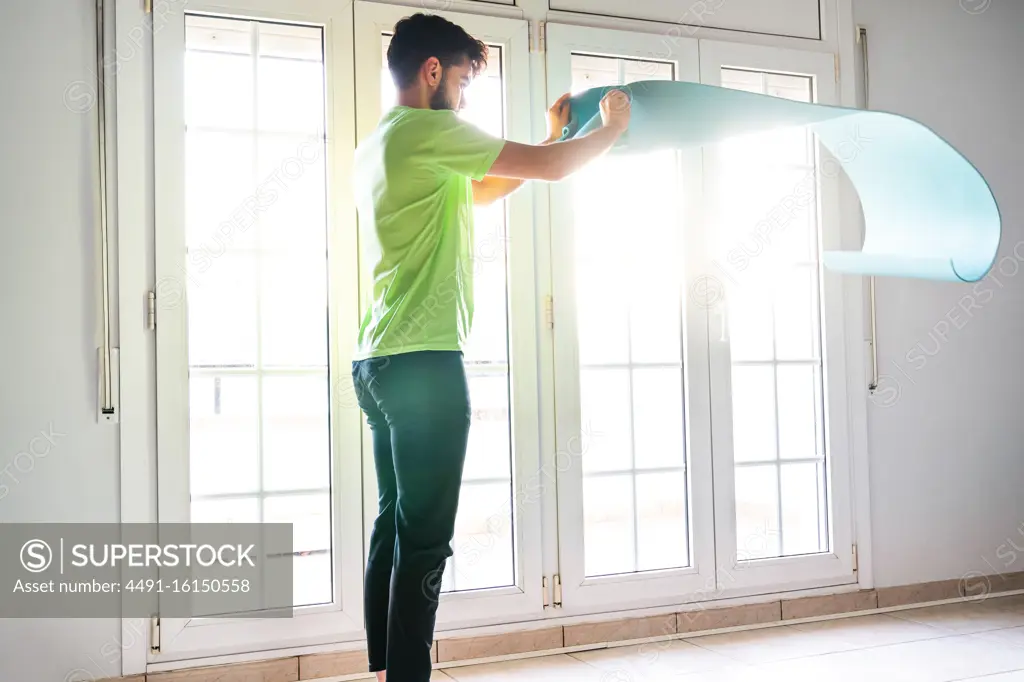 From below back view of tranquil barefoot sportsman in sportswear twisting vivid blue sports mat while finishing workout in light spacious gym