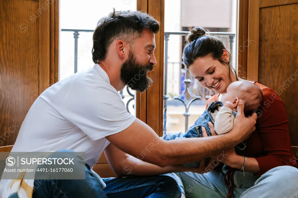 Bearded father and happy mother communicating with cute baby while sitting on floor near window at home