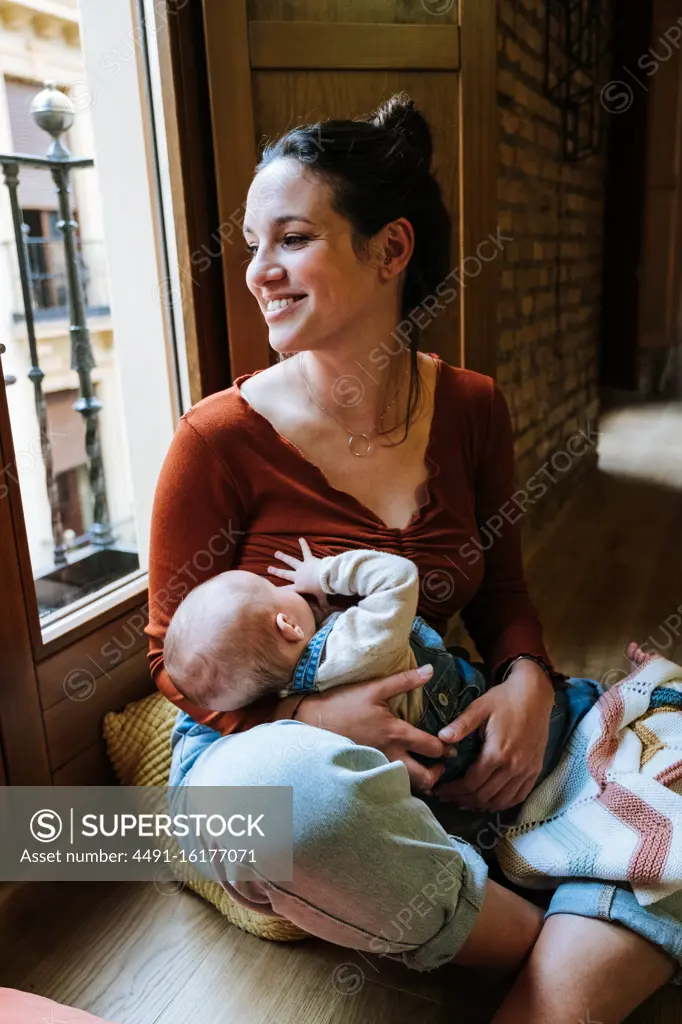 Happy mother breastfeeding baby near window