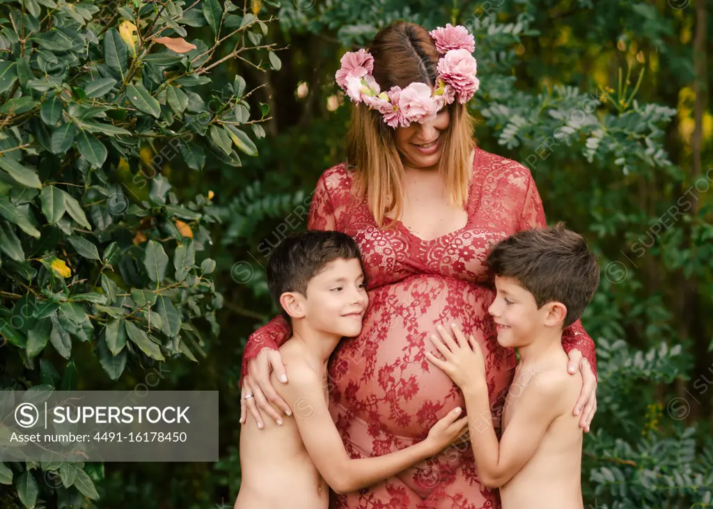 Happy mother in beautiful red dress and floral wreath smiling and embracing twin sons cuddling belly while standing near green trees in garden in summer day