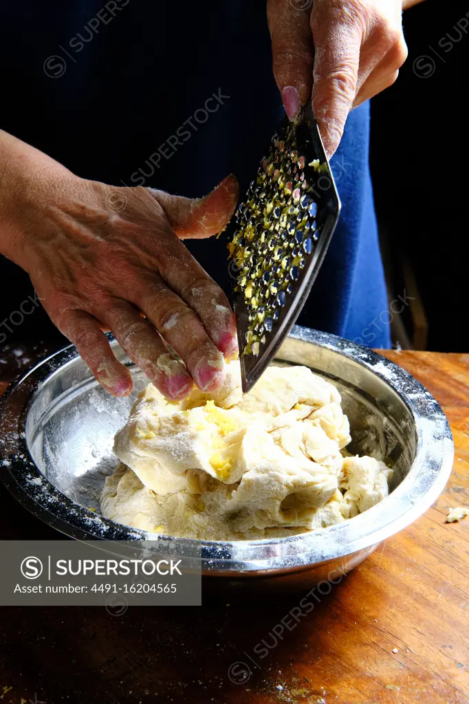 Unrecognizable female removing fresh lemon peel from metal grater over bowl with pastry dough in kitchen