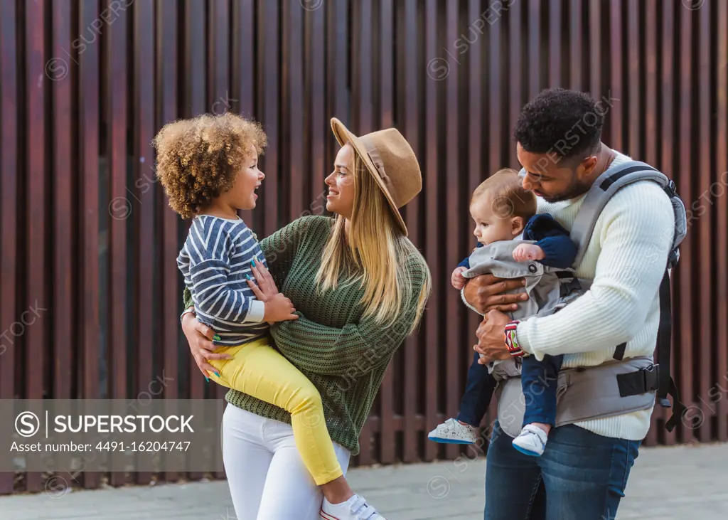 Cheerful stylish young multiracial couple with infant son and little daughter in casual wear walking together along wooden fence on city street