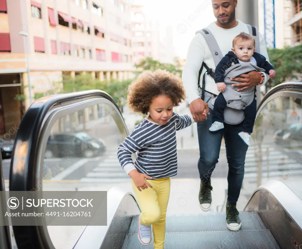 From above of positive young ethnic man carrying infant son and holding hand of cheerful little daughter while standing on escalator in city