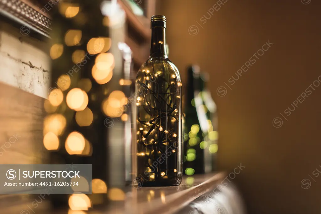 Composed transparent glass bottles with sparkling light garlands inside placed in row on wooden shelf near wall