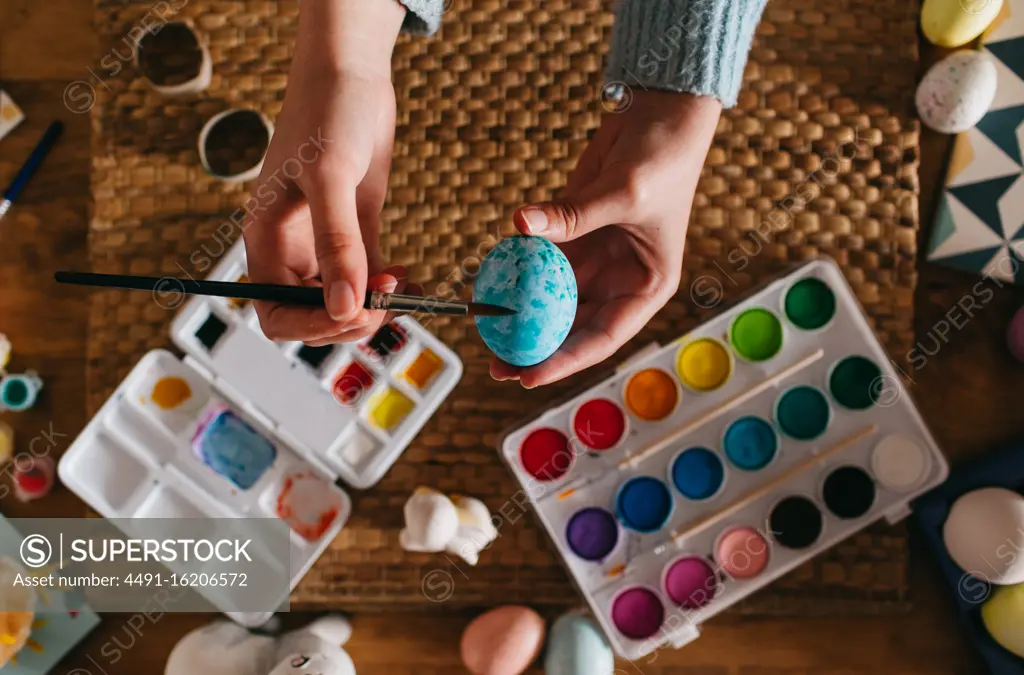 From above top view of cropped unrecognizable person in sweater covering fresh chicken eggs with blue paint while preparing for Easter celebration