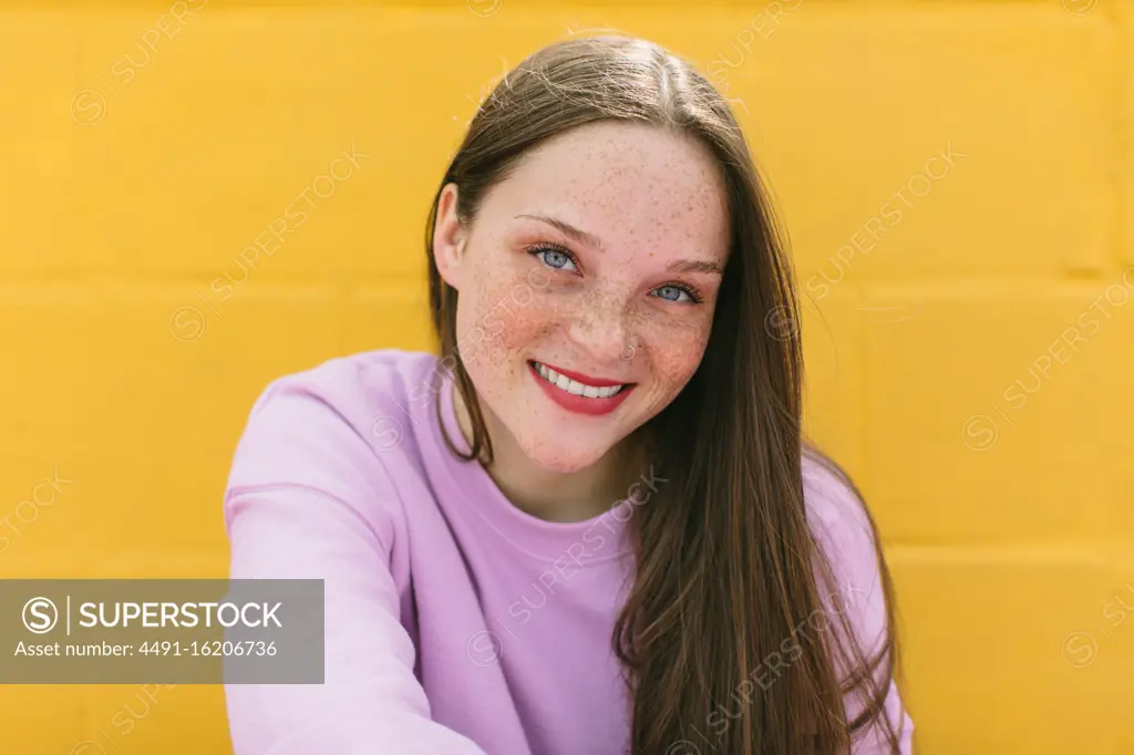 Happy millennial female with freckles in casual wear smiling and looking at camera while sitting against bright yellow stone wall on street