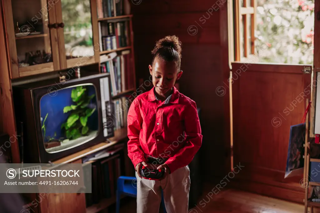 Positive black teen girl in casual clothes with joystick controller in hands playing in room with wooden interior and old fashioned tv