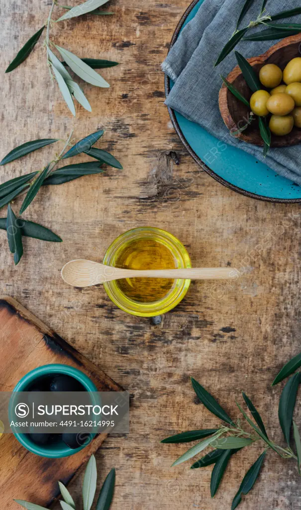 From above of glass jar with organic olive oil with wooden spoon placed on shabby gray table near fresh green olives and tree branches