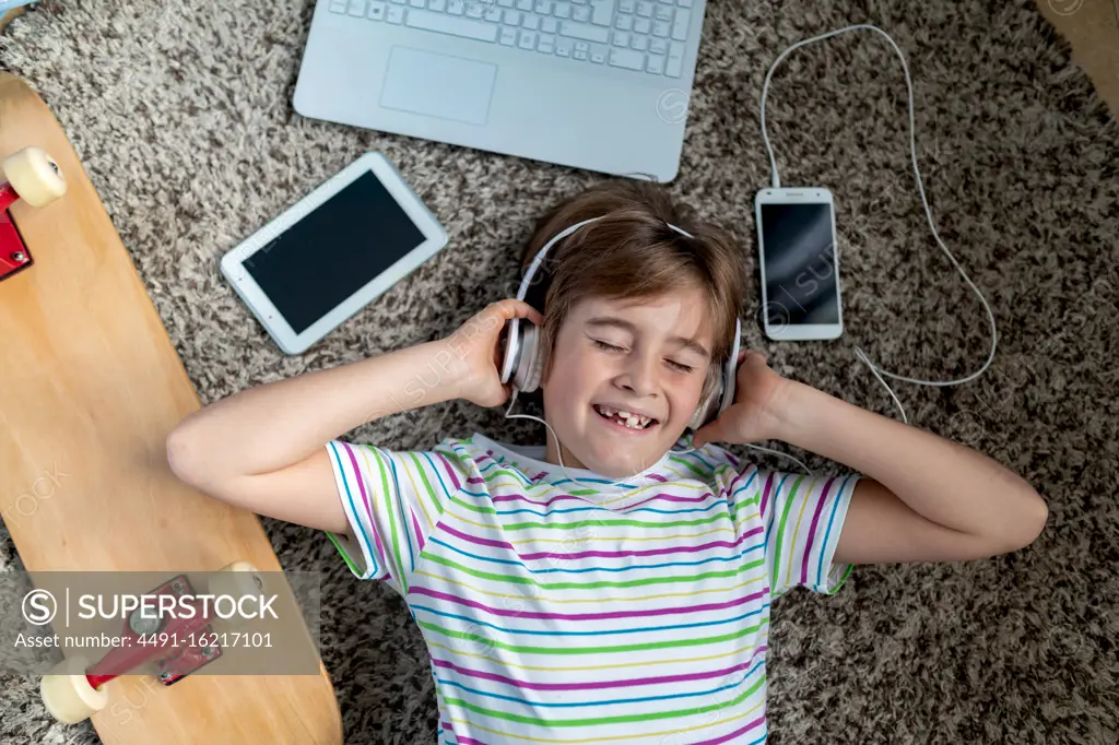 From above of cheerful little boy in casual shirt listening to music with headphones while lying on carpet near gadgets and skateboard in bedroom