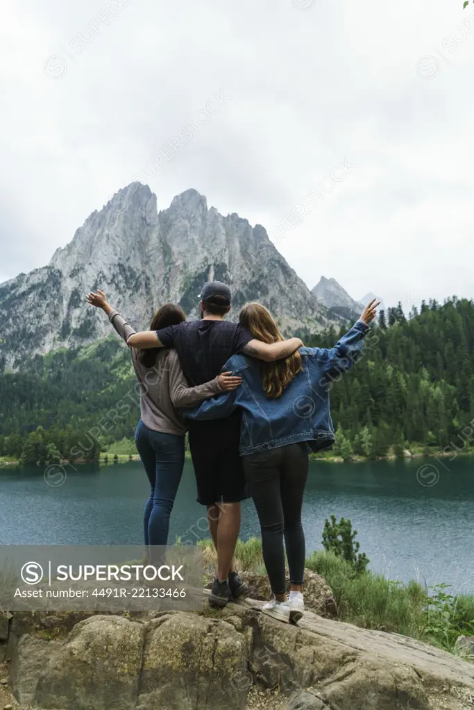 Back view of two women and man embracing and having fun at lake in mountains together.