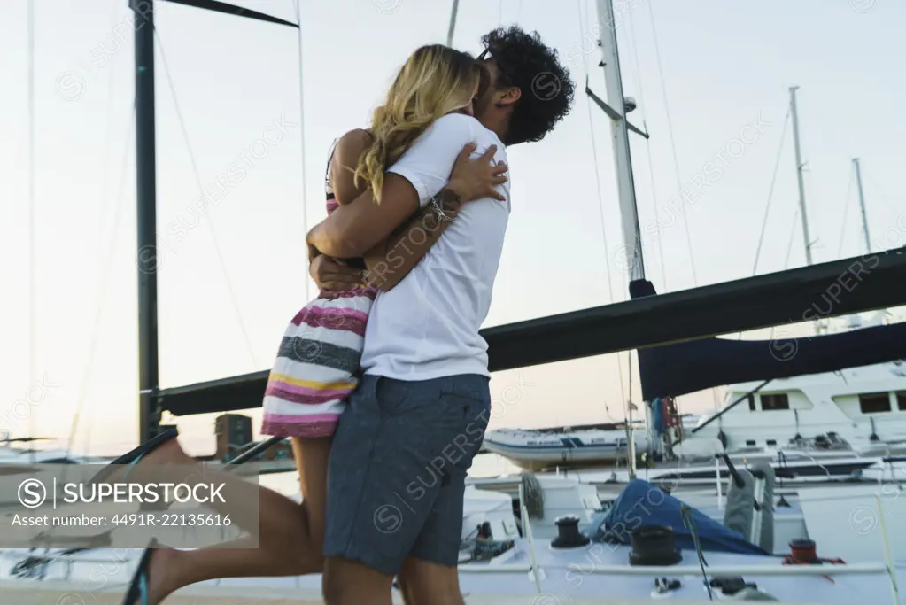 Side view of young loving couple embracing on pier with yachts on background.