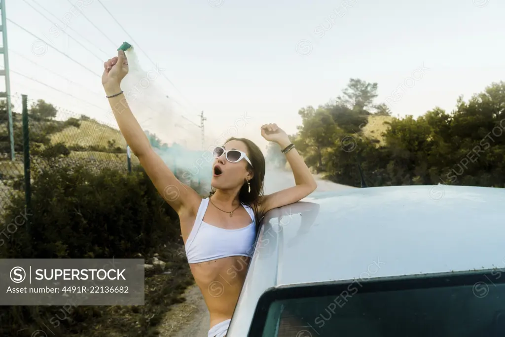 Young woman in sunglasses looking out of car with smoke bomb and shouting happily.