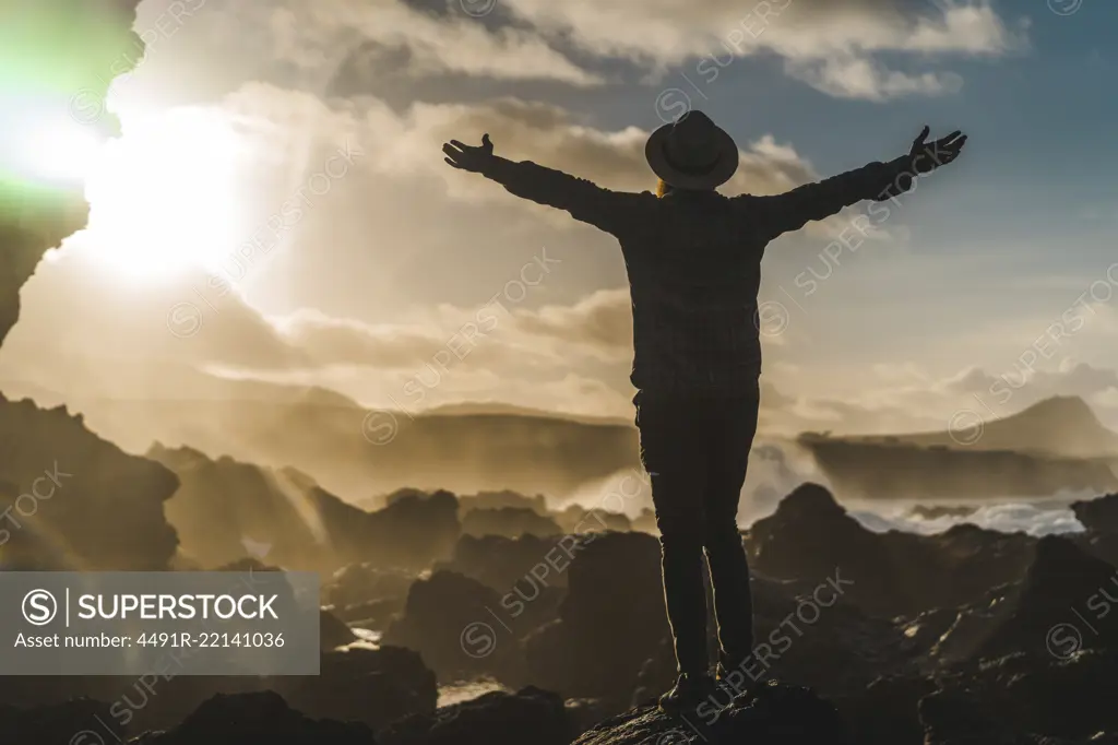 Side view of unrecognizable tourist in hat standing on cliff in sunset lights.