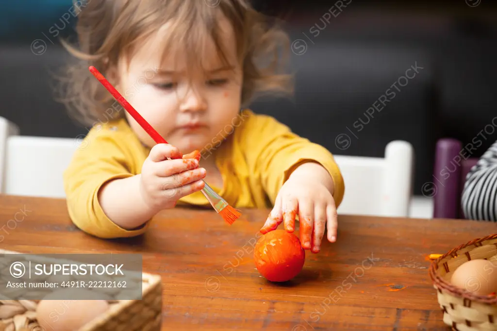 Adorable toddler girl using paintbrush to paint Easter egg while sitting at table