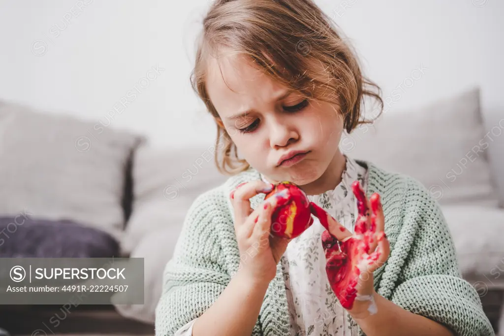 Funny child with hands in colors painting Easter egg near sofa on blurred background