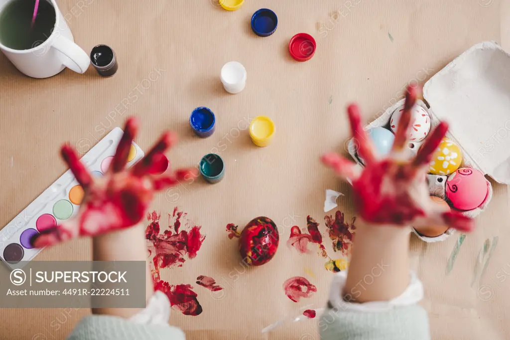 From above crop palms of kid in paints near Easter eggs in container and set of bright colors on carton background