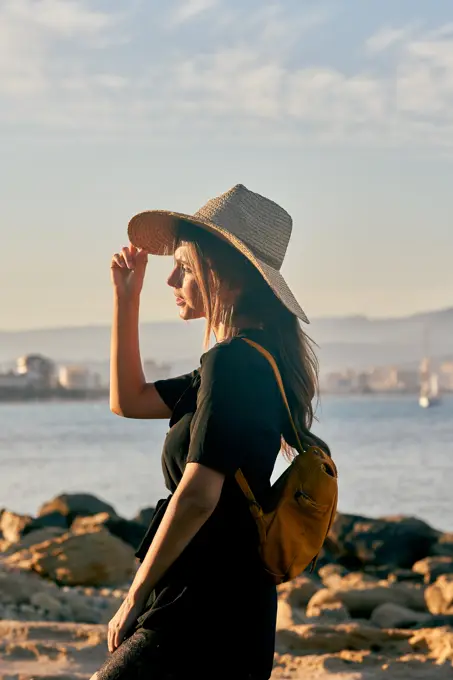 Side view of female tourist wearing a straw hat and backpack standing near beach