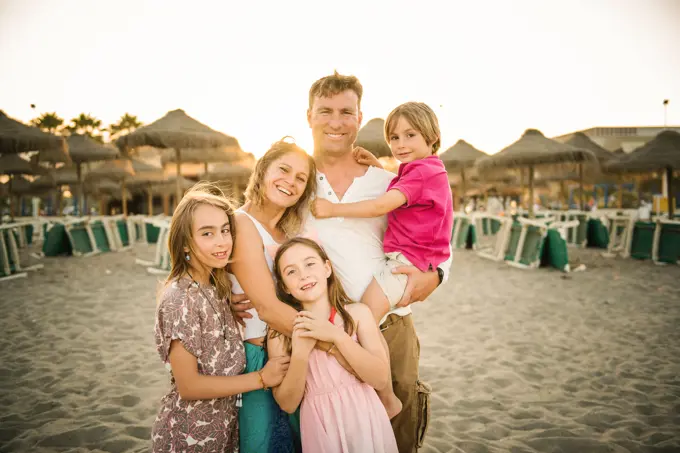 Adult loving man and woman with son and daughters standing together on beach in back lit smiling at camera