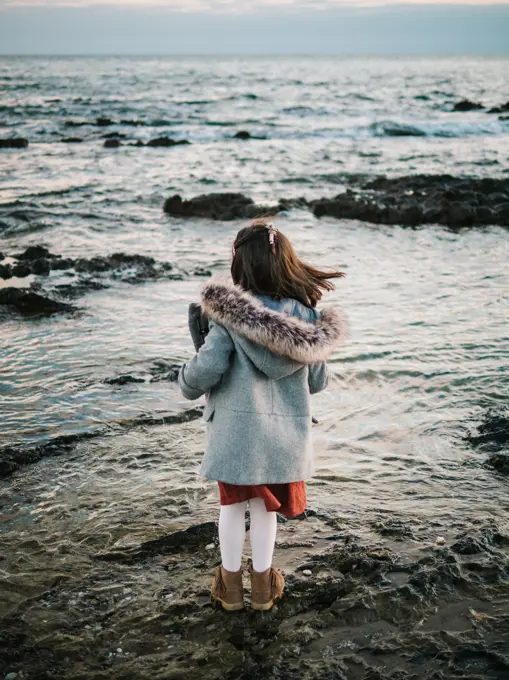 anonymous little girl from behind standing by the beach on rocks looking at the sea on a winter day