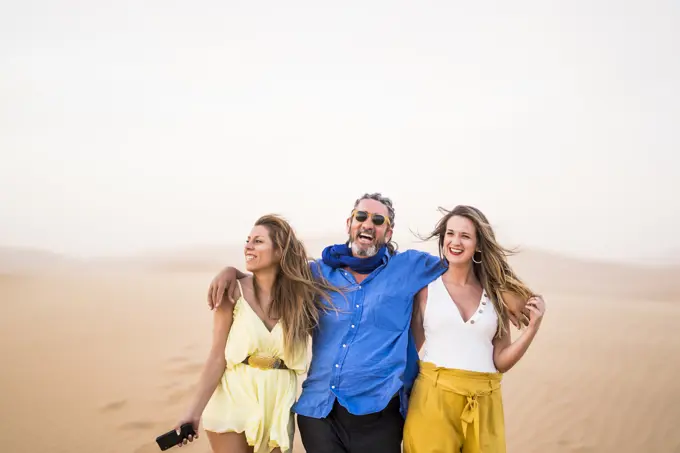 Senior bearded man laughing and hugging cheerful women while walking in sandy desert during trip in Morocco