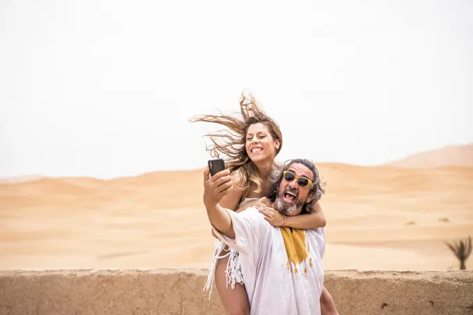 Middle-aged man with woman piggyback taking selfie expressively on terrace against sandy desert, Morocco