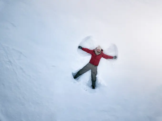 From above smiling middle-aged male in outwear lying on white snow and making snow angel silhouette