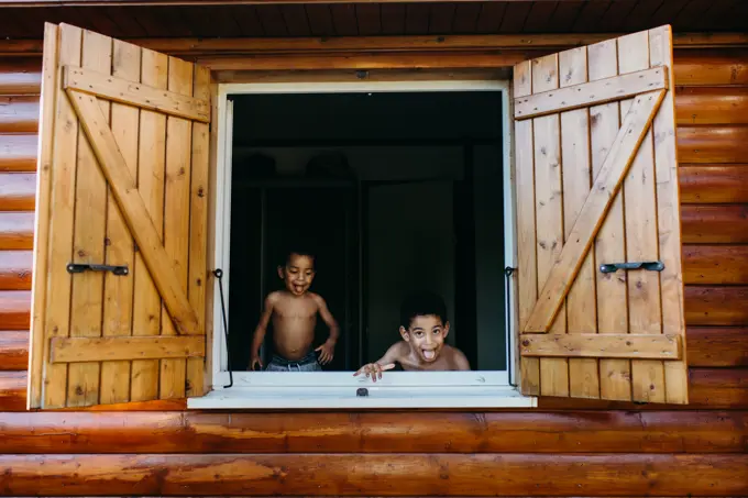 Two shirtless African American brothers looking out open window of wooden house while having fun at home together