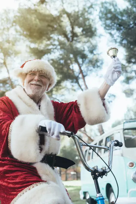 From below senior man in costume of Santa Claus sitting on cycle, ringing bell and looking at camera