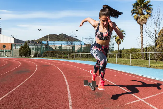 Strong female athlete in sportswear running fast against blue sky on sunny day on stadium