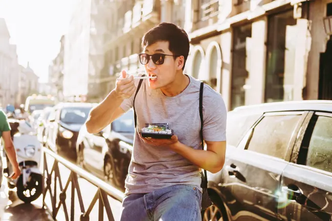 Ethnic handsome asian man in sunglasses eating takeaway food with chopsticks while standing by food truck on sunny street