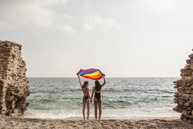Back view of lesbian couple standing on beach holding hands and waving colorful flag of LGBT movement during summer vacation