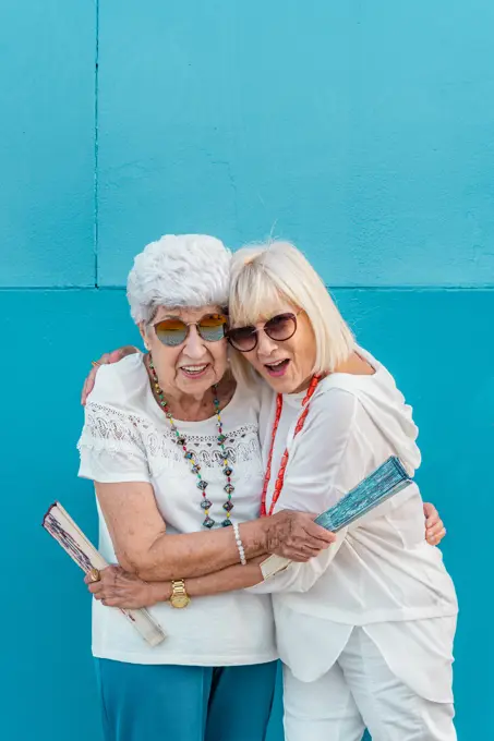 Positive trendy aged grey-haired females waving with big color hand fans talking looking at each other on blue background in summer outside