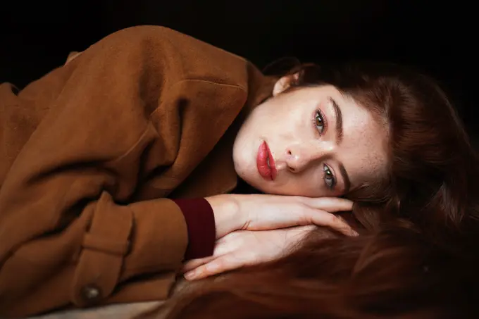 Charming thoughtful woman in brown coat lying on hands dreaming while looking away