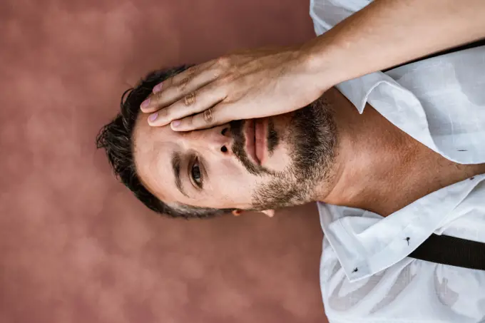 From above classy man in white shirt and suspenders lying on back and floating looking at camera with hand covering an eye in sea at sandbank